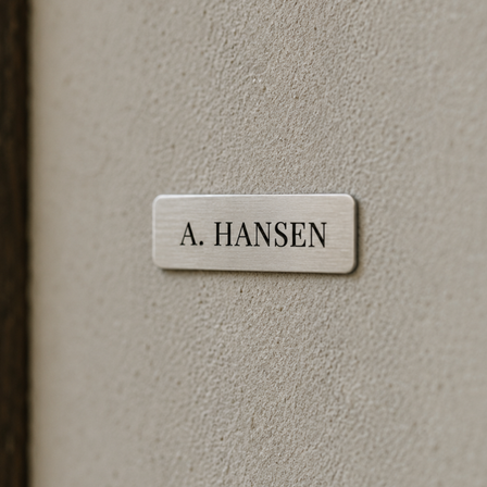 Lille navneskilt i forniklet messing med teksten “A. HANSEN”, monteret på en lys væg ved siden af en mørk trædør