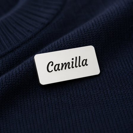 Hvidt navneskilt i graveret plast med sort bundlag og navnet “Camilla”, fotograferet tæt på mørkeblåt finstrikket stof.