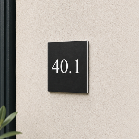 Lille sort skilt i graveret plast med hvid bund og det indgraverede nummer “40.1”, monteret tæt på en lys facade ved siden af en mørk hoveddør.