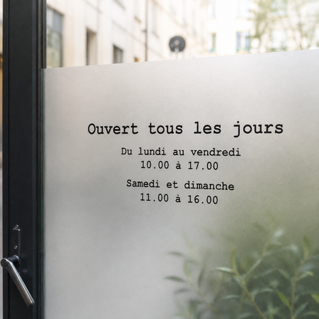 Texte adhésif noir indiquant les horaires d’ouverture, appliqué sur une vitrine en verre dépoli dans un environnement lumineux et naturel.